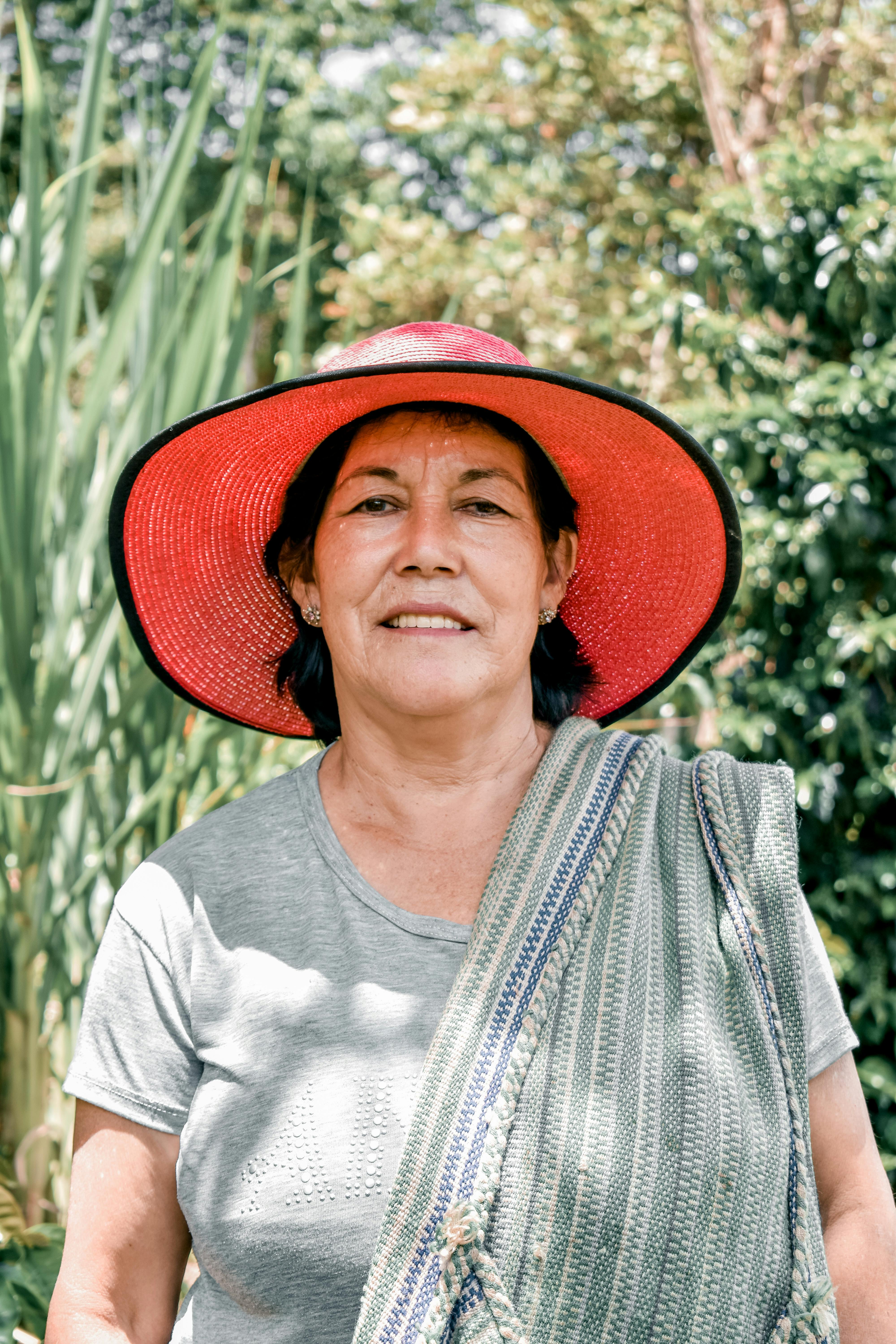 Senior woman smiling in nature wearing a red hat and shawl. Outdoors, vibrant greenery.