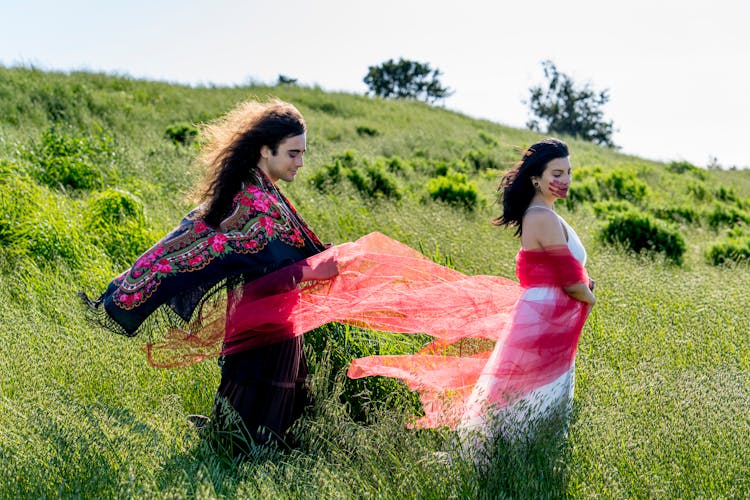 Man With Long Hair And Woman Wearing Dresses And Standing On A Meadow 