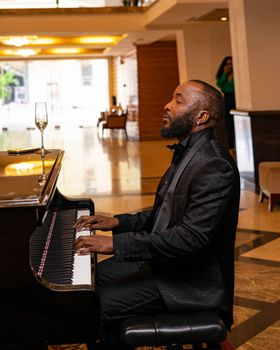 A stylish pianist in a black suit plays gracefully indoors with a champagne flute nearby.