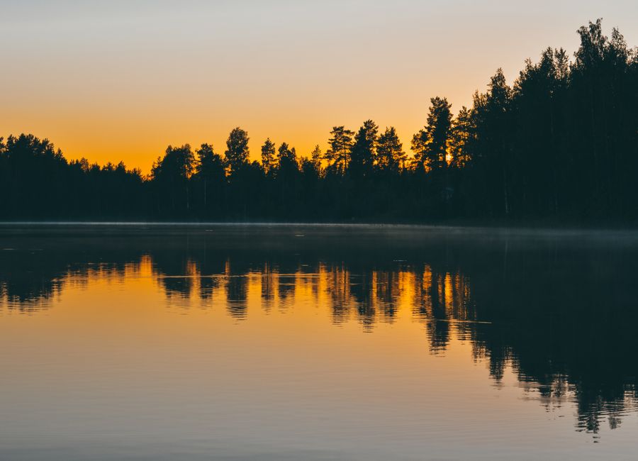 Peaceful early morning sunrise over a tranquil lake in Jyväskylä, Finland, with tree silhouettes reflecting in the water.