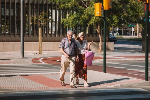 Free A senior couple walks across a sunny street in Málaga, Spain, carrying shopping bags. Stock Photo
