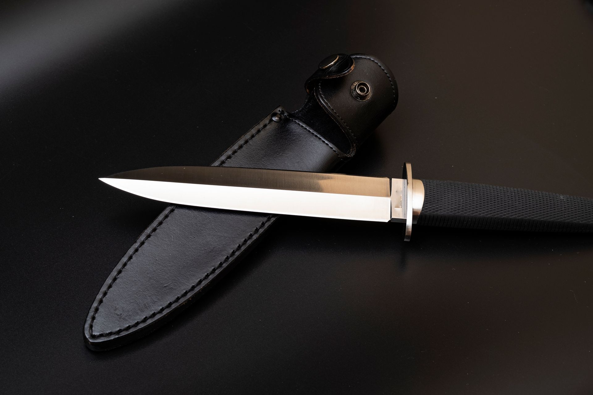 A polished stainless steel dagger and black leather sheath on a dark background.