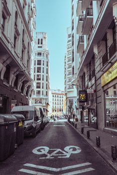 A narrow urban street in Madrid, Spain, lined with buildings and parked cars on a sunny day.