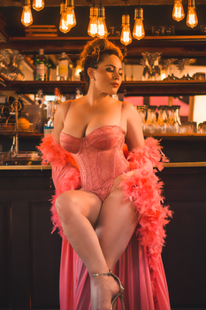 A woman in pink lingerie sits elegantly at a bar under warm lighting.