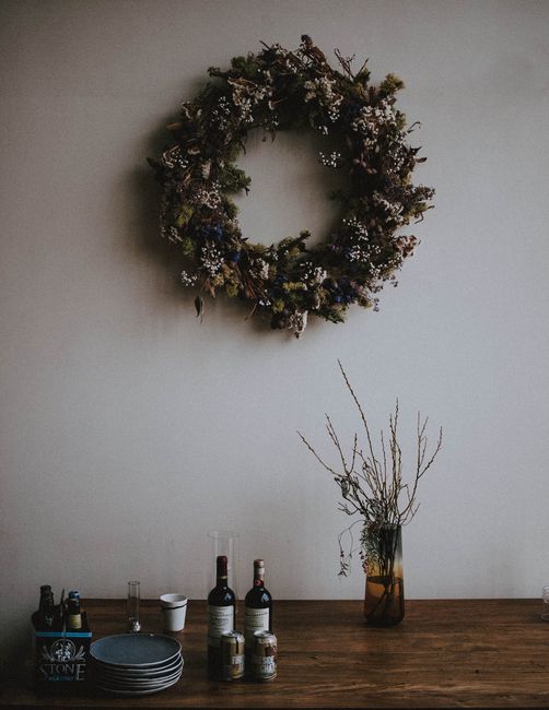 A rustic wreath hangs above a wooden table with bottles and dry flowers.