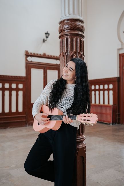 Woman with long black hair smiling and playing a guitar against a wooden column indoors.