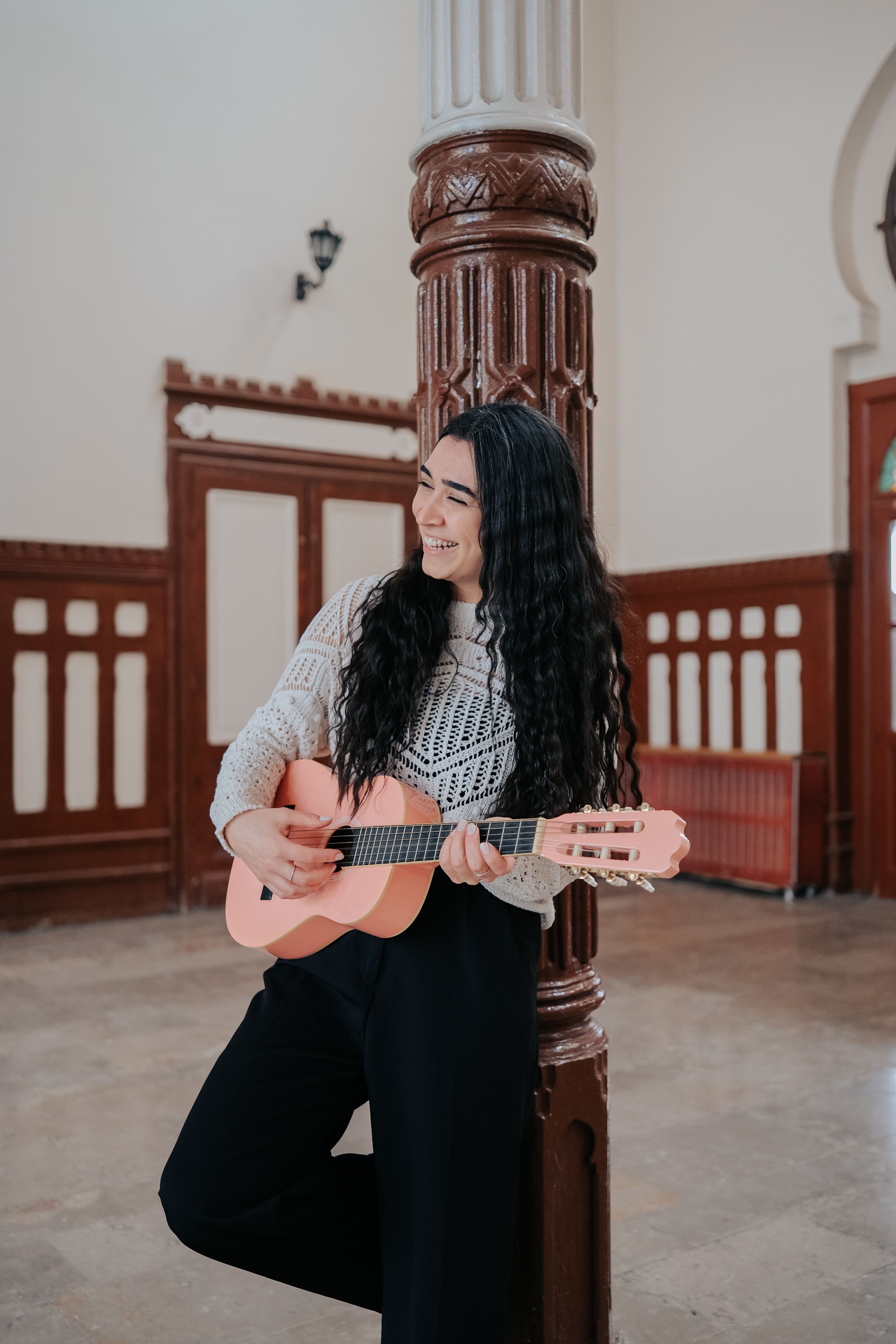 A young woman stands indoors playing a ukulele with a joyful smile, leaning against a column.
