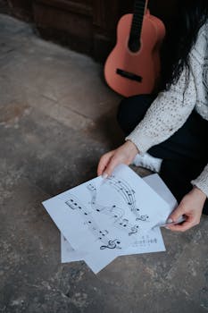 A woman sitting indoors organizing musical sheets on the floor with a guitar in the background.