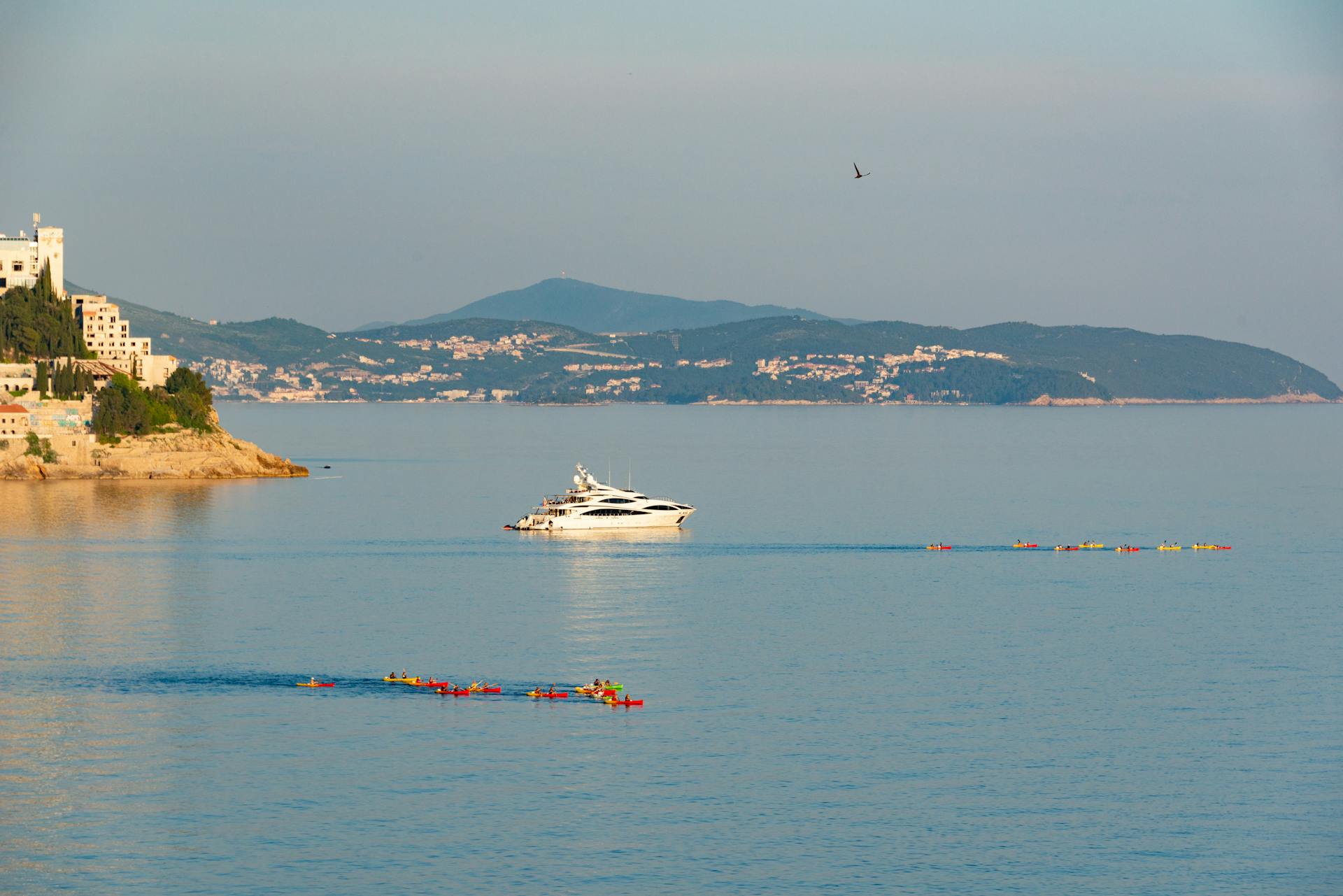 Scenic view of a luxury yacht and kayakers by the Dubrovnik coastline, perfect for summer travel and tourism.
