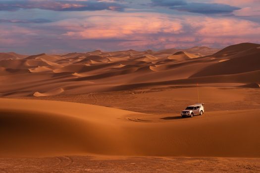 A vehicle traverses vast desert dunes under a beautiful sunset sky, showcasing the rugged landscape.