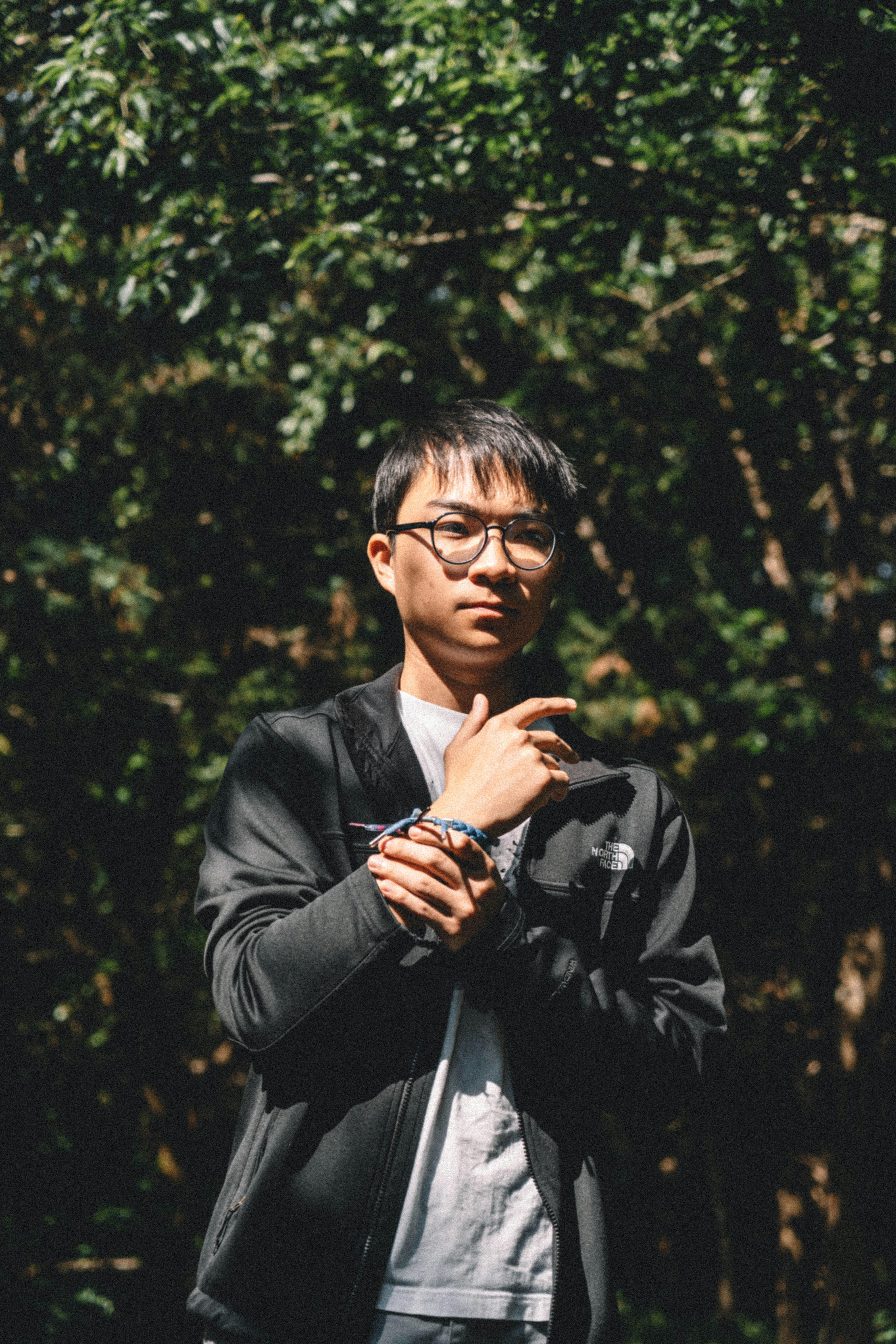 Asian man wearing glasses and a bracelet standing in a sunlit forest setting.