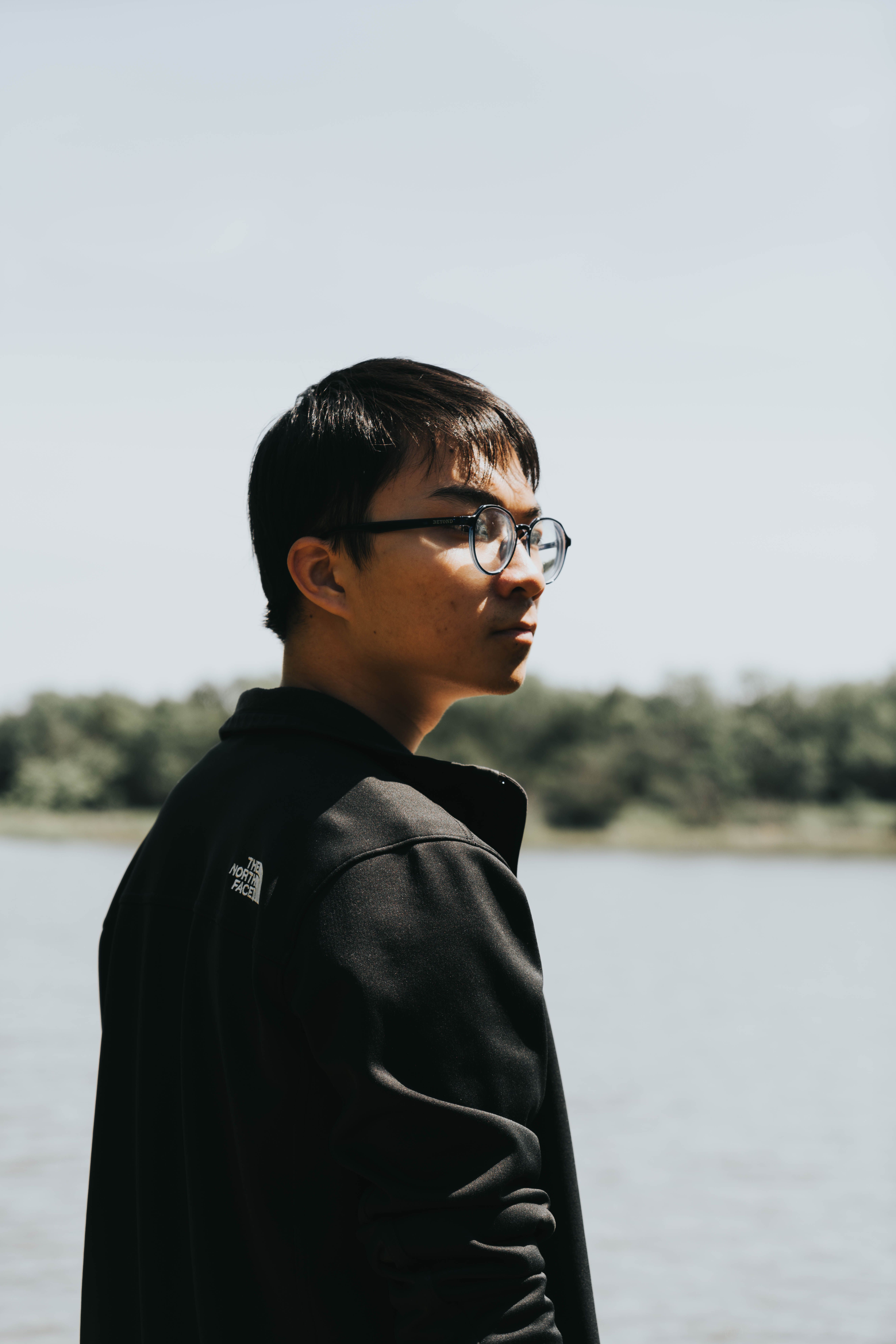 A young man with glasses and black hair gazes into the distance by a lakeside in a black jacket.