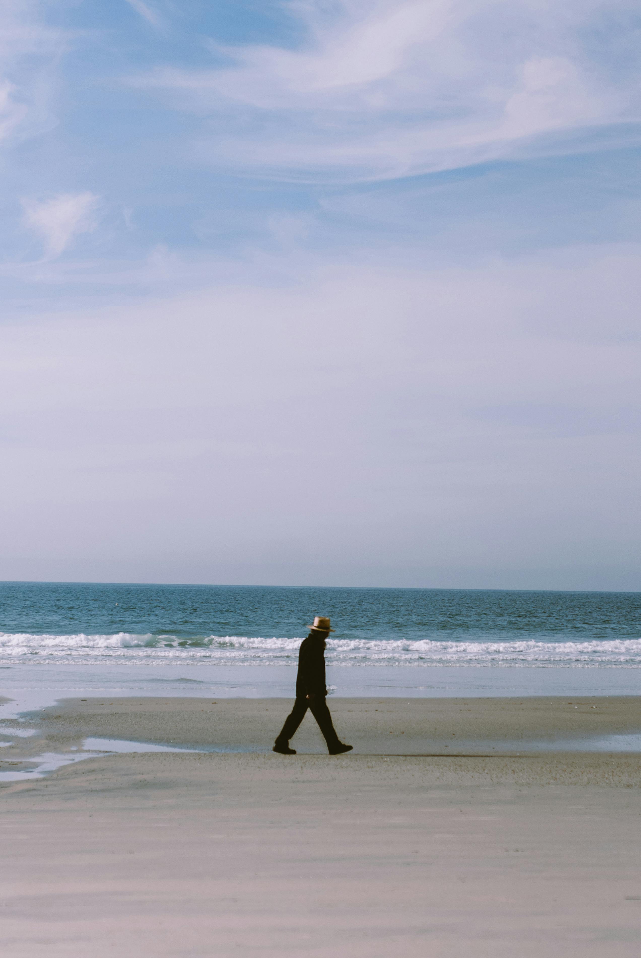 A solitary man in a hat walks on a serene beach with waves in Tijuana, Mexico.