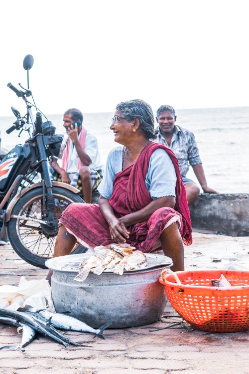 Free An elderly woman selling fish outdoors by the sea, sitting beside her catch and a motorbike. Stock Photo