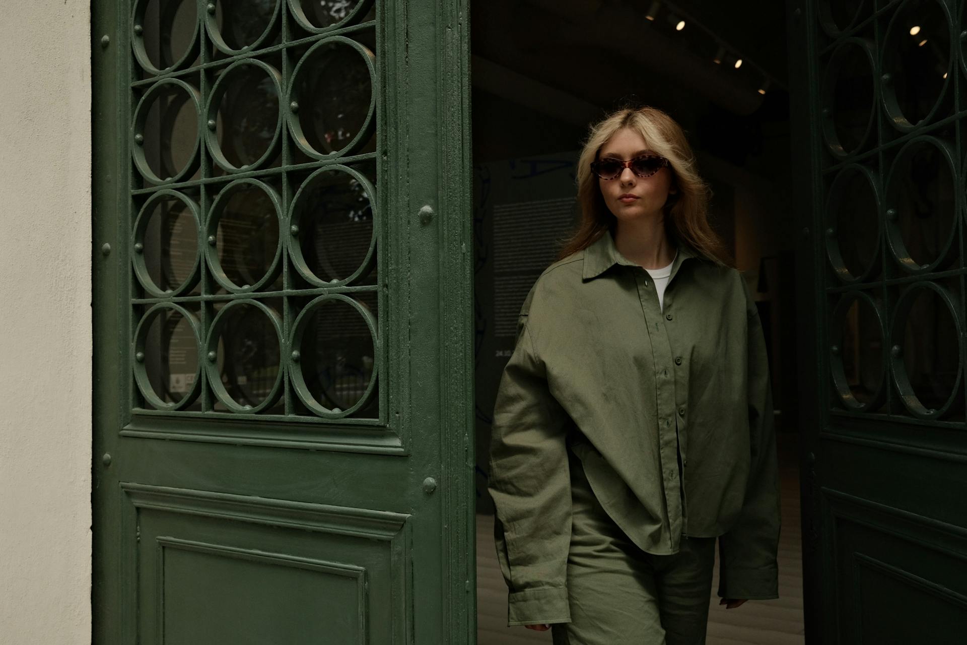 Woman in olive green oversized outfit and sunglasses exiting a vintage doorway.