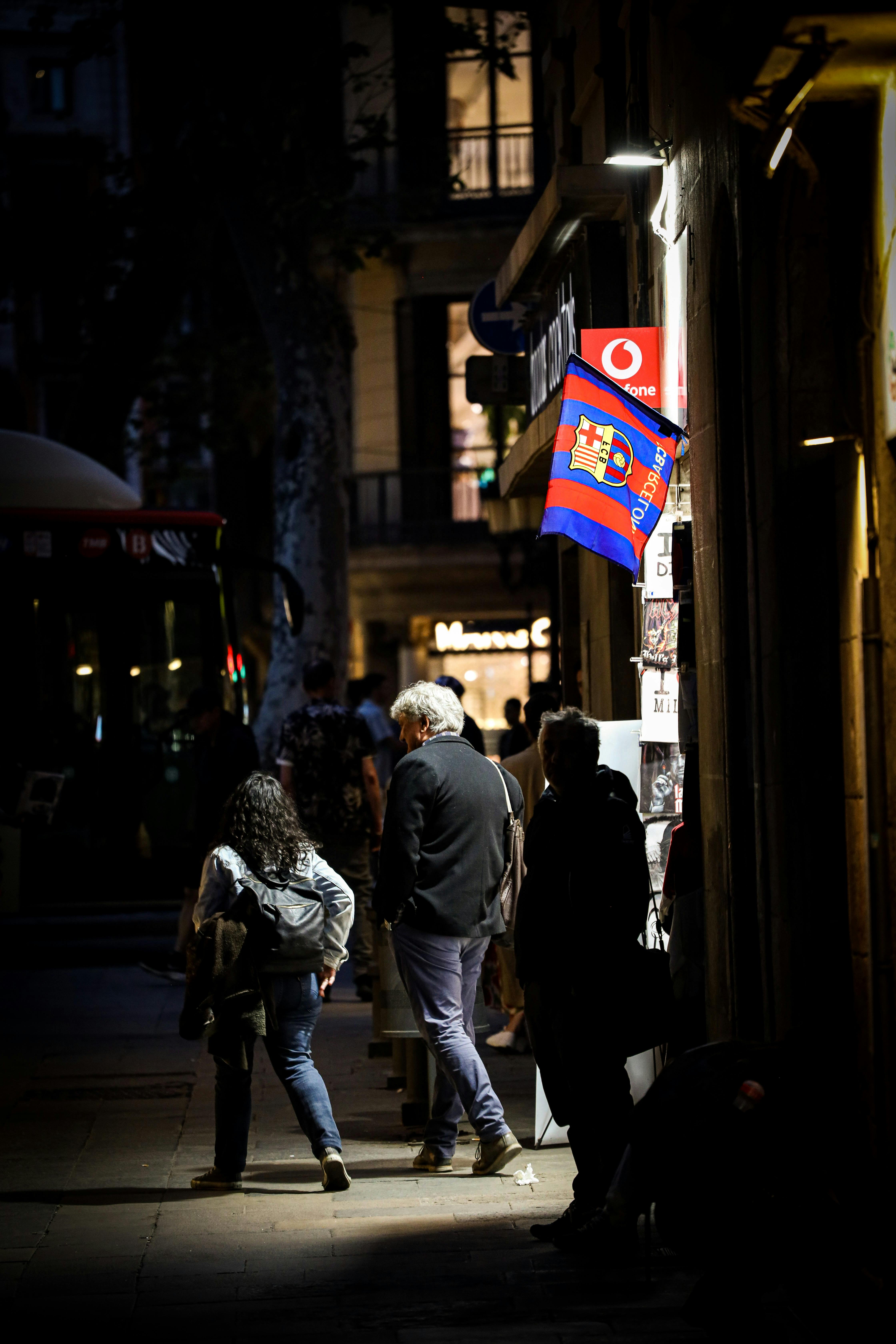 people walking on a dark street with barcelona football club flag on a building