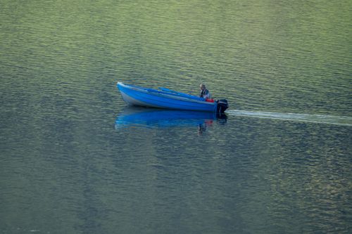 Free A lone blue boat cruises quietly on a serene lake, reflecting verdant surroundings. Stock Photo