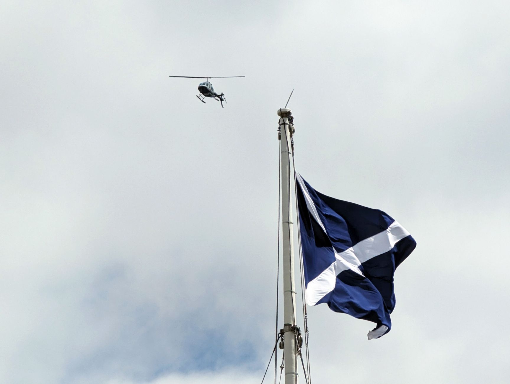 A helicopter flies over a Scottish flag waving on a flagpole against a cloudy sky.