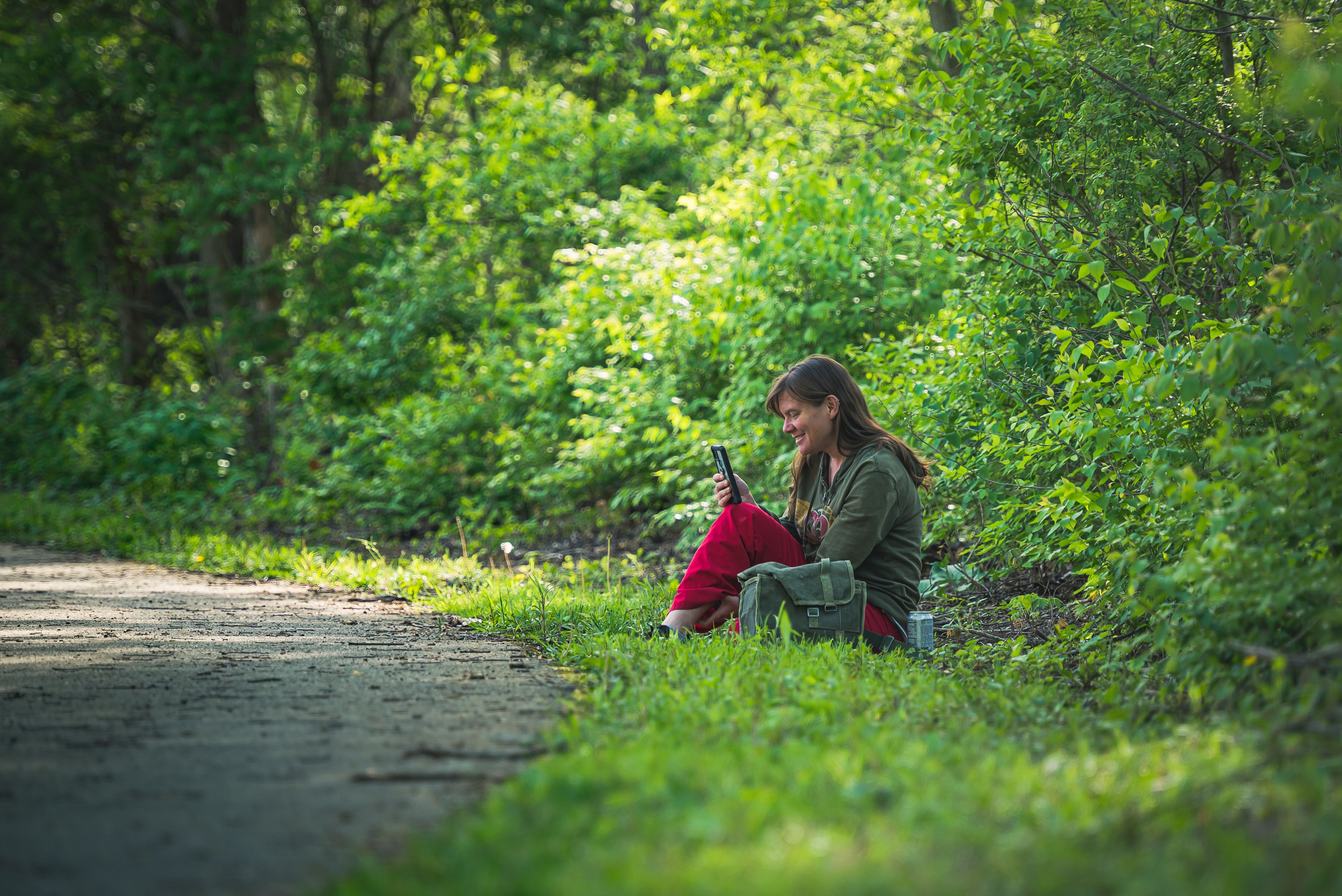 Woman sitting on a dirt road edge, using a smartphone amidst lush greenery.
