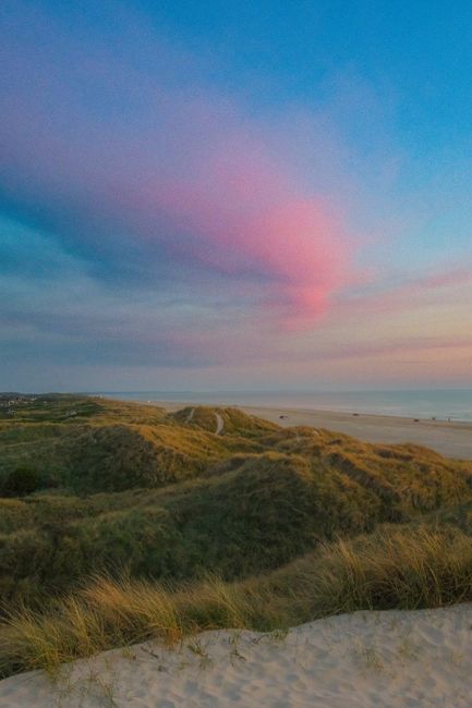 A serene view of grassy coastal dunes against a vibrant, colorful sunset sky over the ocean horizon.