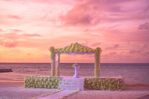 White Table Under Brown Concrete Gazebo Along Body of Water Under White Orange and Pink Cloudy Sky during Sunset