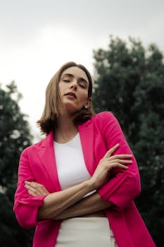 A stylish woman wearing a pink blazer stands with arms crossed outdoors.