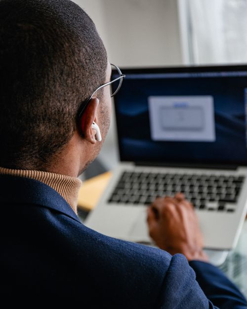 Free Back view of a man using a laptop with earbuds, focusing on his task indoors. Stock Photo