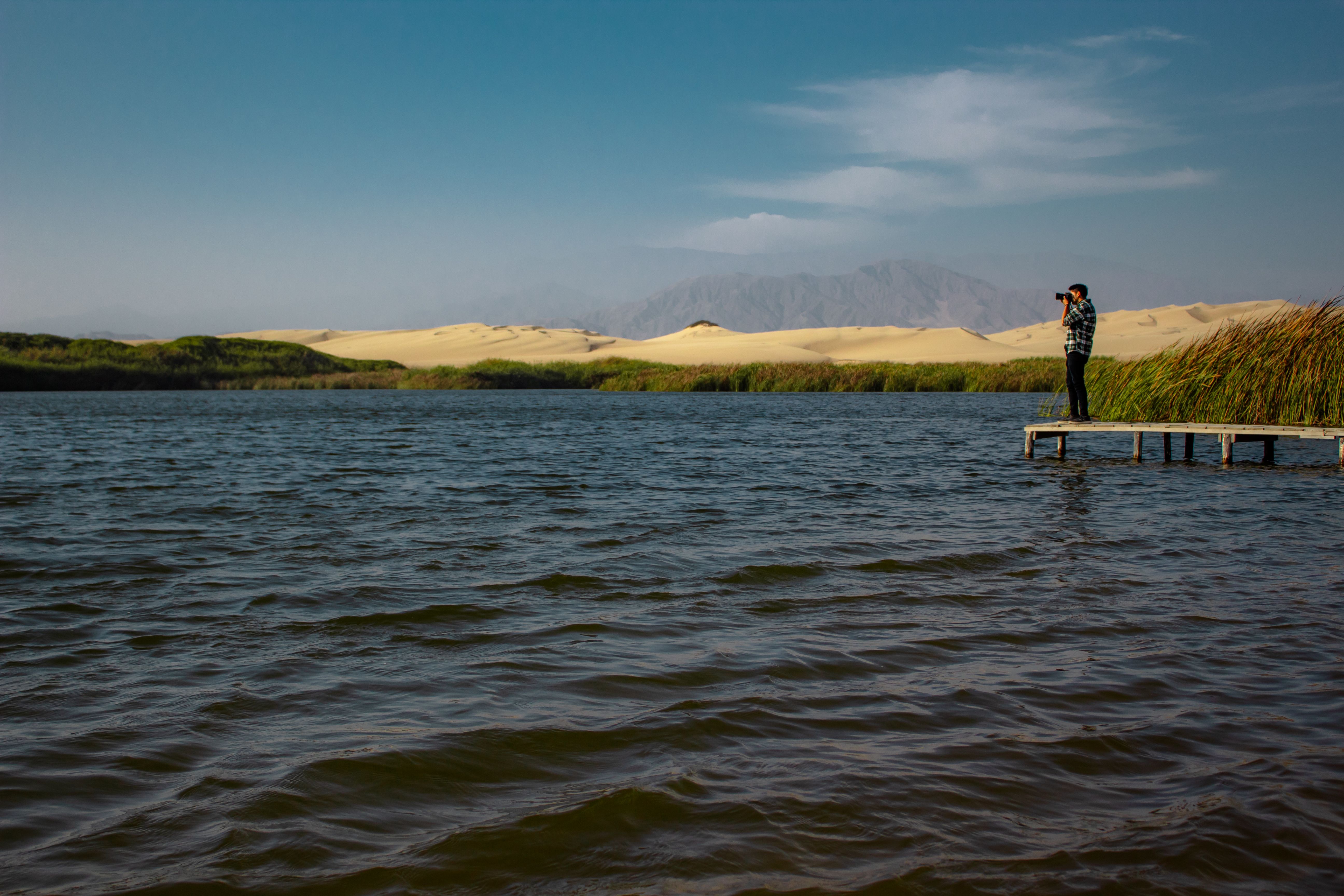 A lone photographer stands on a dock, capturing desert lakeside scenery in Pisco, Peru.