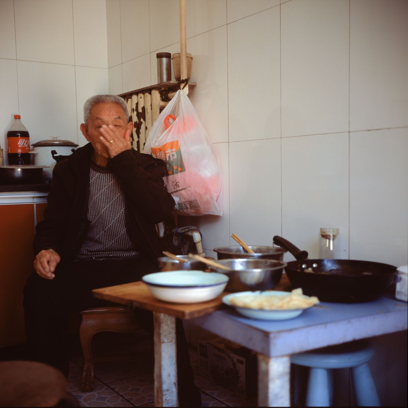 Portrait of an elderly man sitting in a kitchen with cooking tools and dishes on the table.