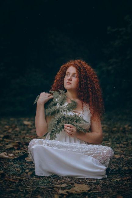 A red-haired woman in a white dress holding fern leaves in a serene forest setting.