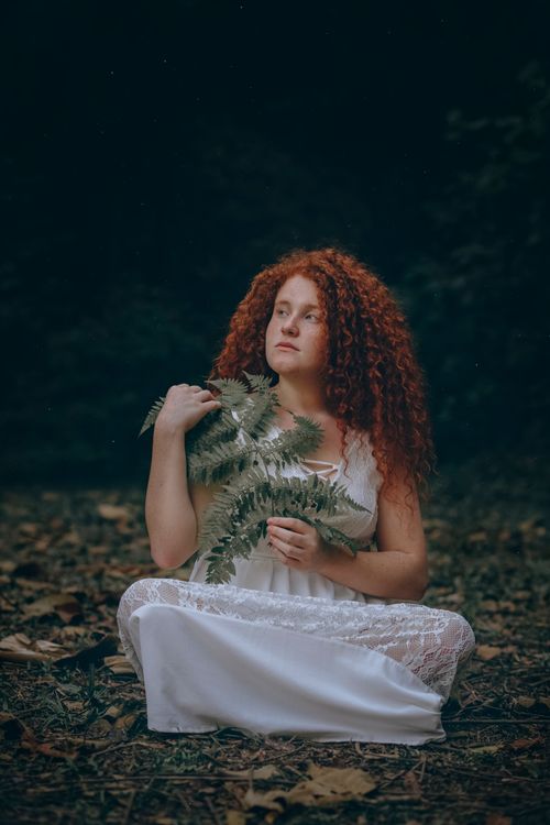 Free A red-haired woman in a white dress holding fern leaves in a serene forest setting. Stock Photo
