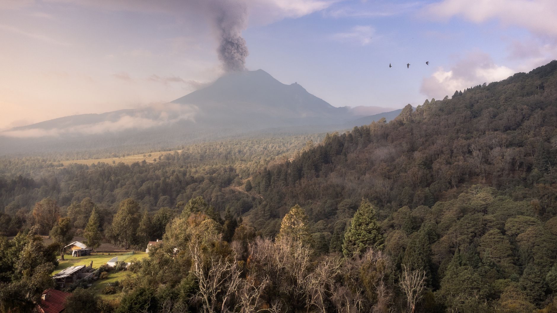 Dramatic aerial shot of a smoking volcano in Mexico with surrounding dense forest and rural scenery.