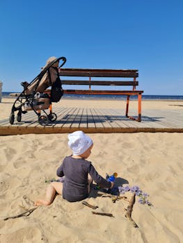 A baby plays on a sandy beach with a stroller and bench, perfect for summer vacation themes.