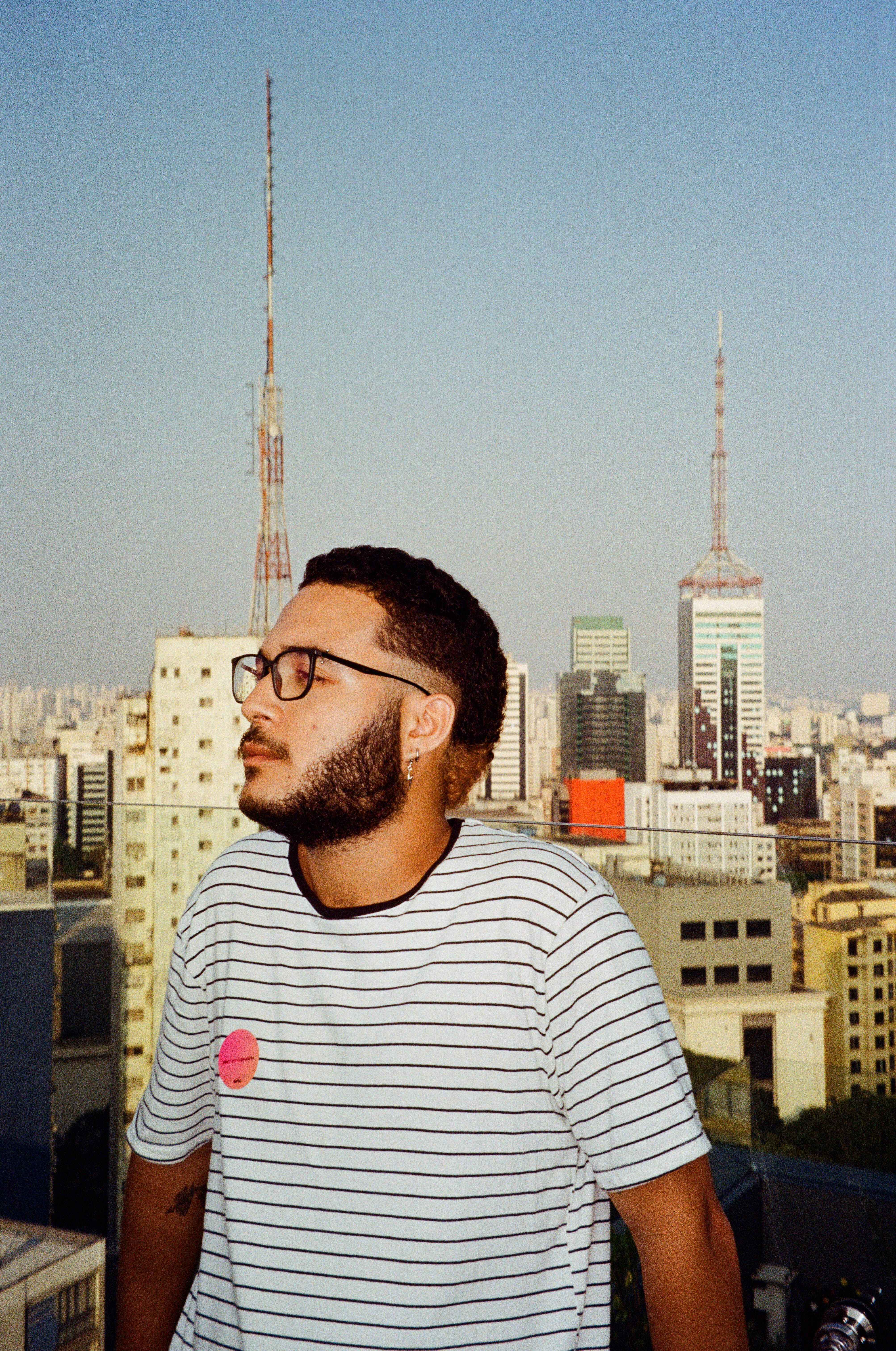 A young man with glasses poses against São Paulo's distinctive skyline on a sunny day.