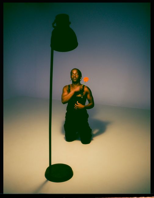 A man kneeling indoors holding a flower under dramatic lighting, creating a moody artistic atmosphere.