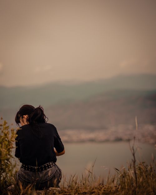 Free A young woman sits peacefully on a grassy hill overlooking a serene lake at sunset. Stock Photo