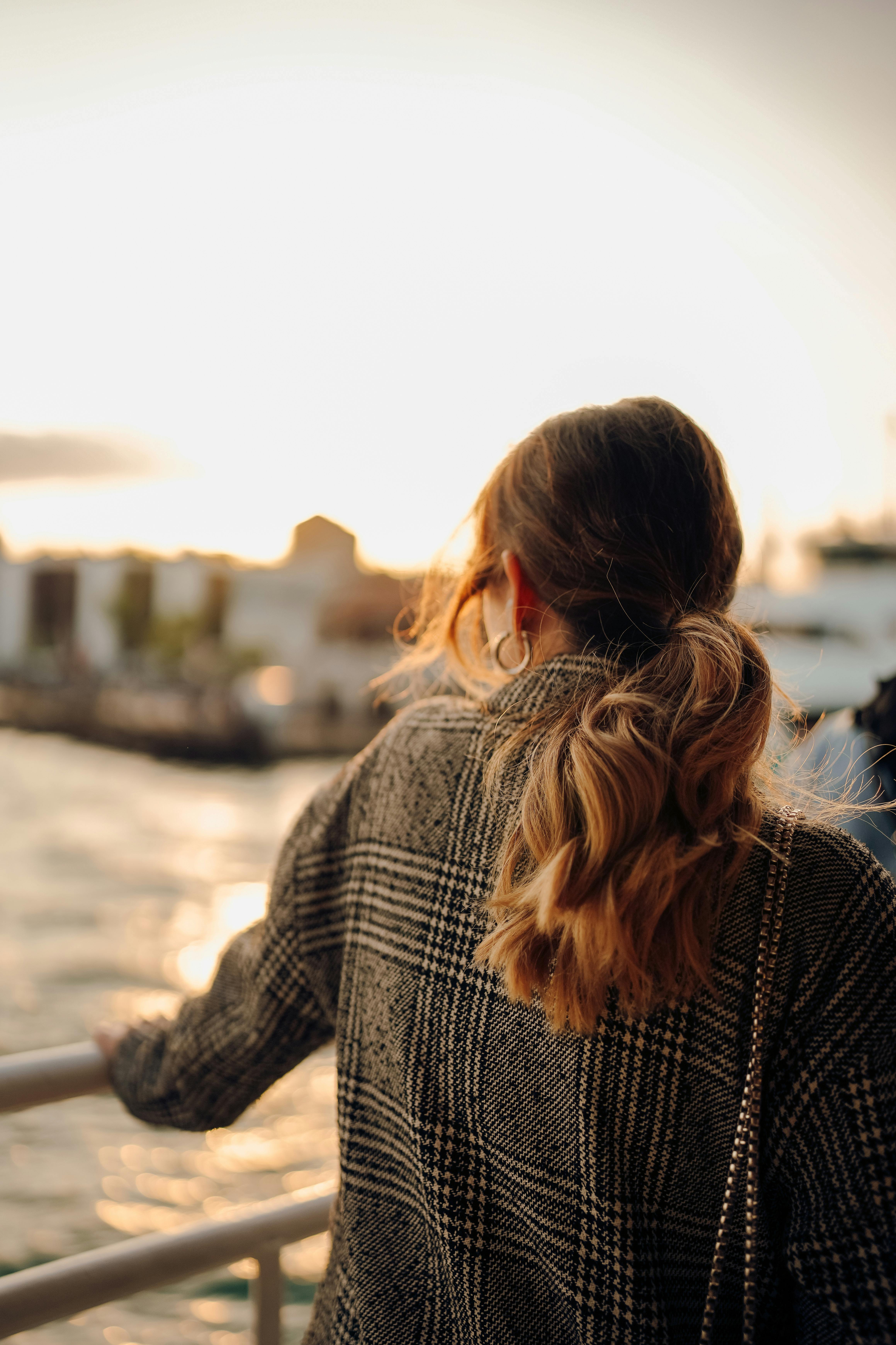 A woman with a ponytail leans on a railing, enjoying a sea view at sunset.