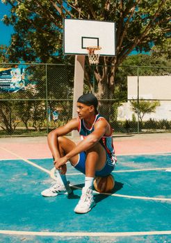 Athlete in sportswear sitting on a basketball at an outdoor court during the day.