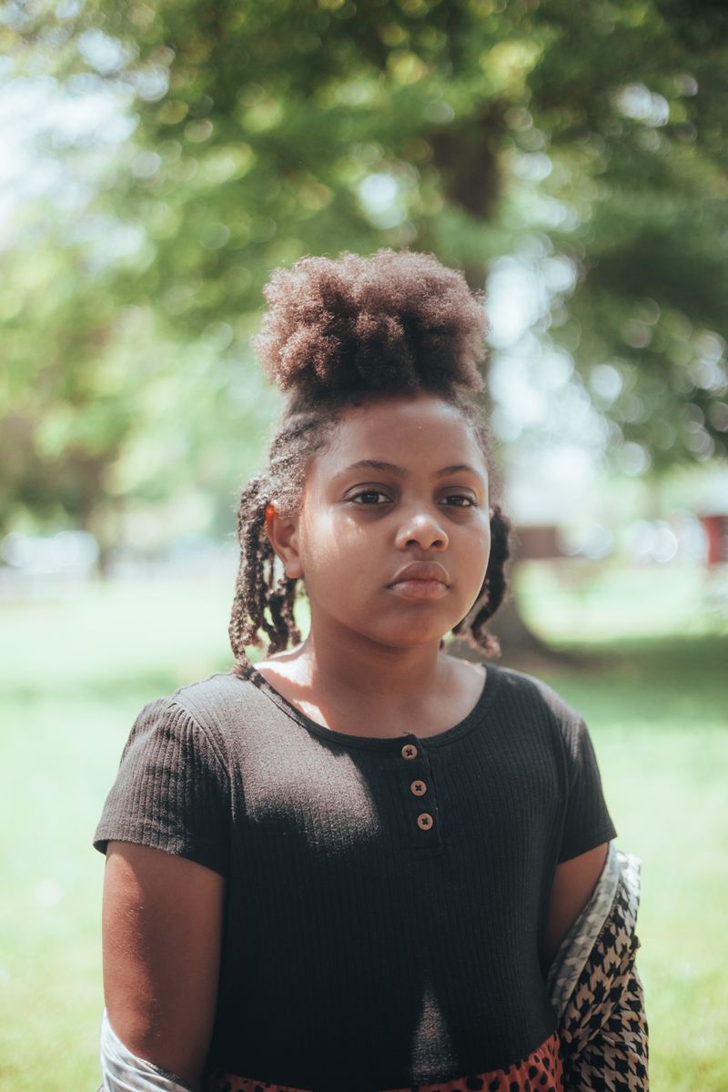 A Black teenage girl with natural hair in an updo stands outdoors in a park, the age when the HPV vaccine is routinely recommended.