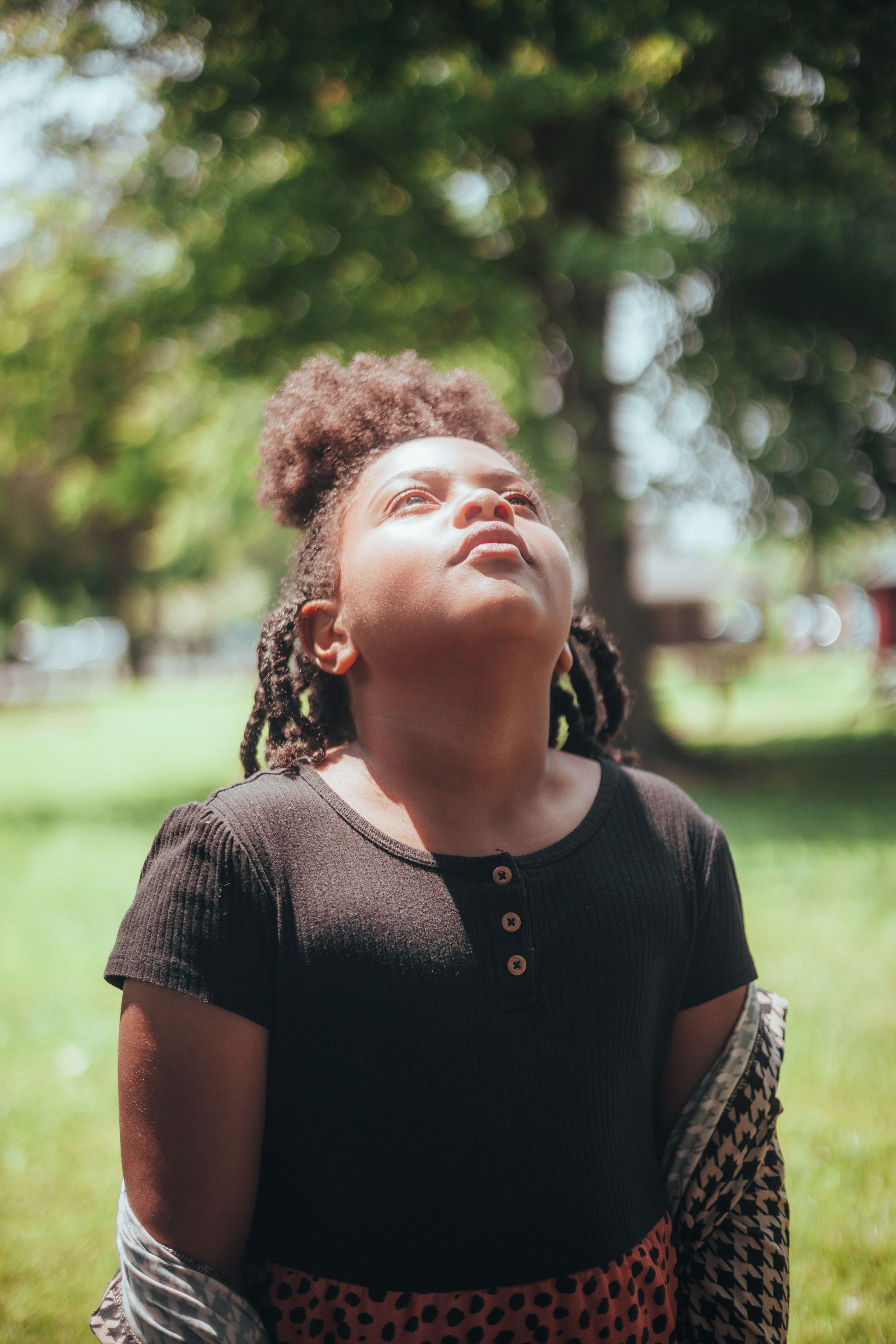 A young girl in a park looking upwards, surrounded by lush green trees under bright sunlight