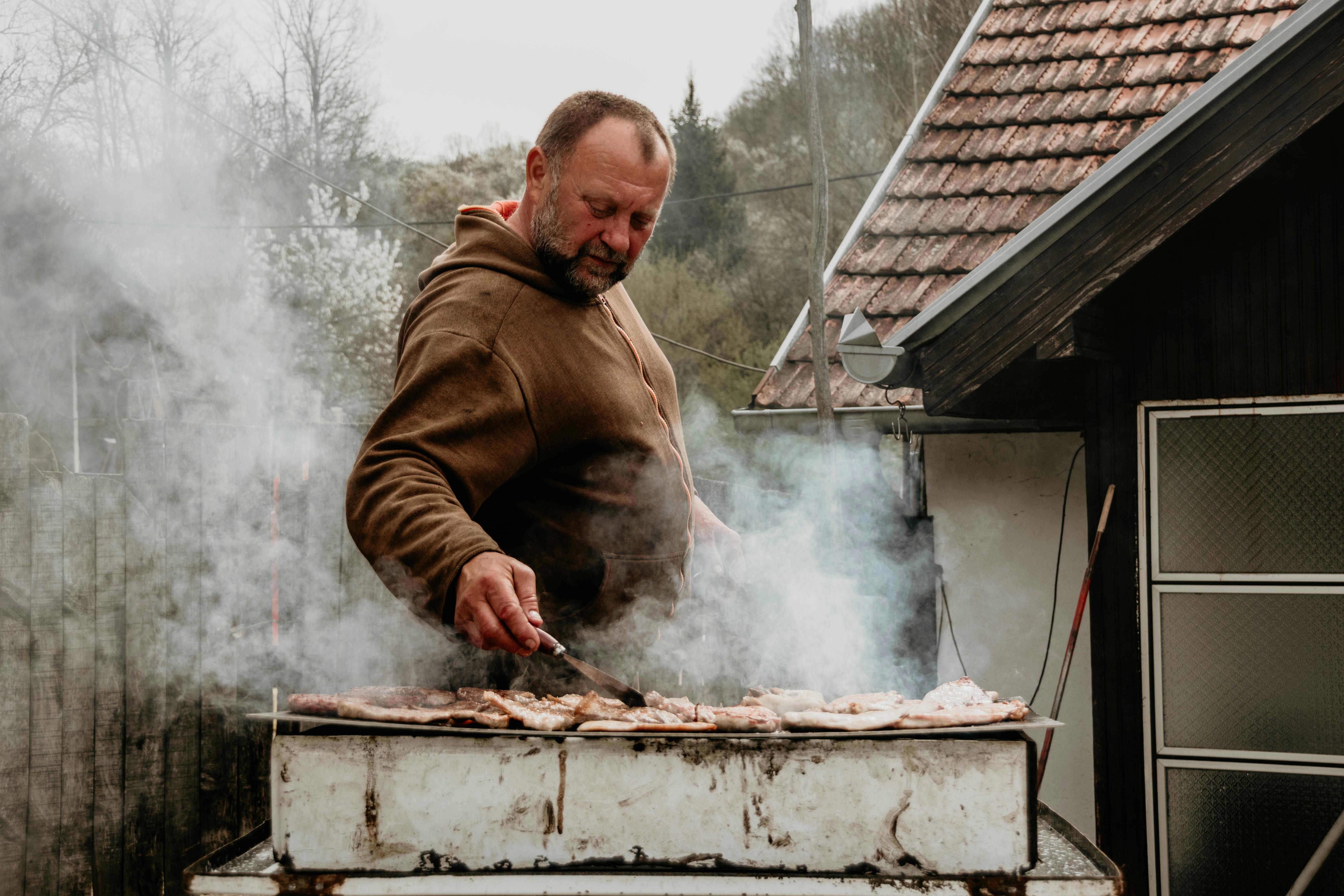 A man grills barbecue outdoors in rural Belgrade, Serbia, surrounded by smoke and nature.