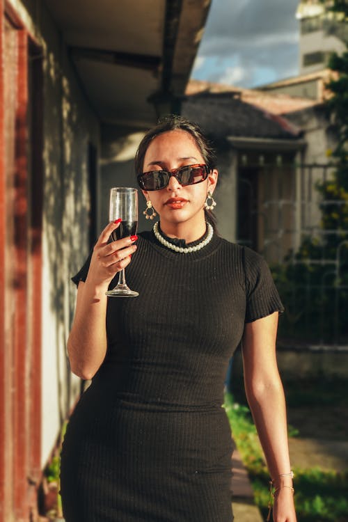 Young Woman in a Black Dress and Sunglasses Standing Outdoors with a Drink in Hand 