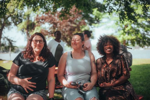 Three diverse women enjoying a sunny day outdoors, seated on a bench and smiling.