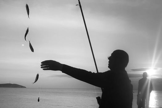 Silhouetted man fishing with multiple catches dangling at sunset by the sea.