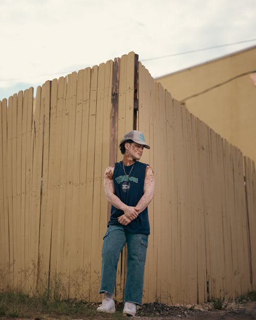 Man in Cap and Tank Top Standing in Corner of Wooden Fence