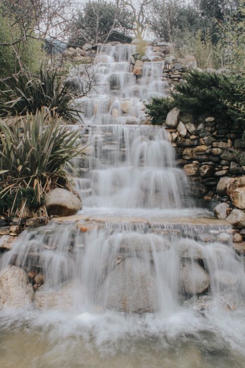 Free A beautiful waterfall flows over stones surrounded by lush green plants, creating a tranquil scene. Stock Photo