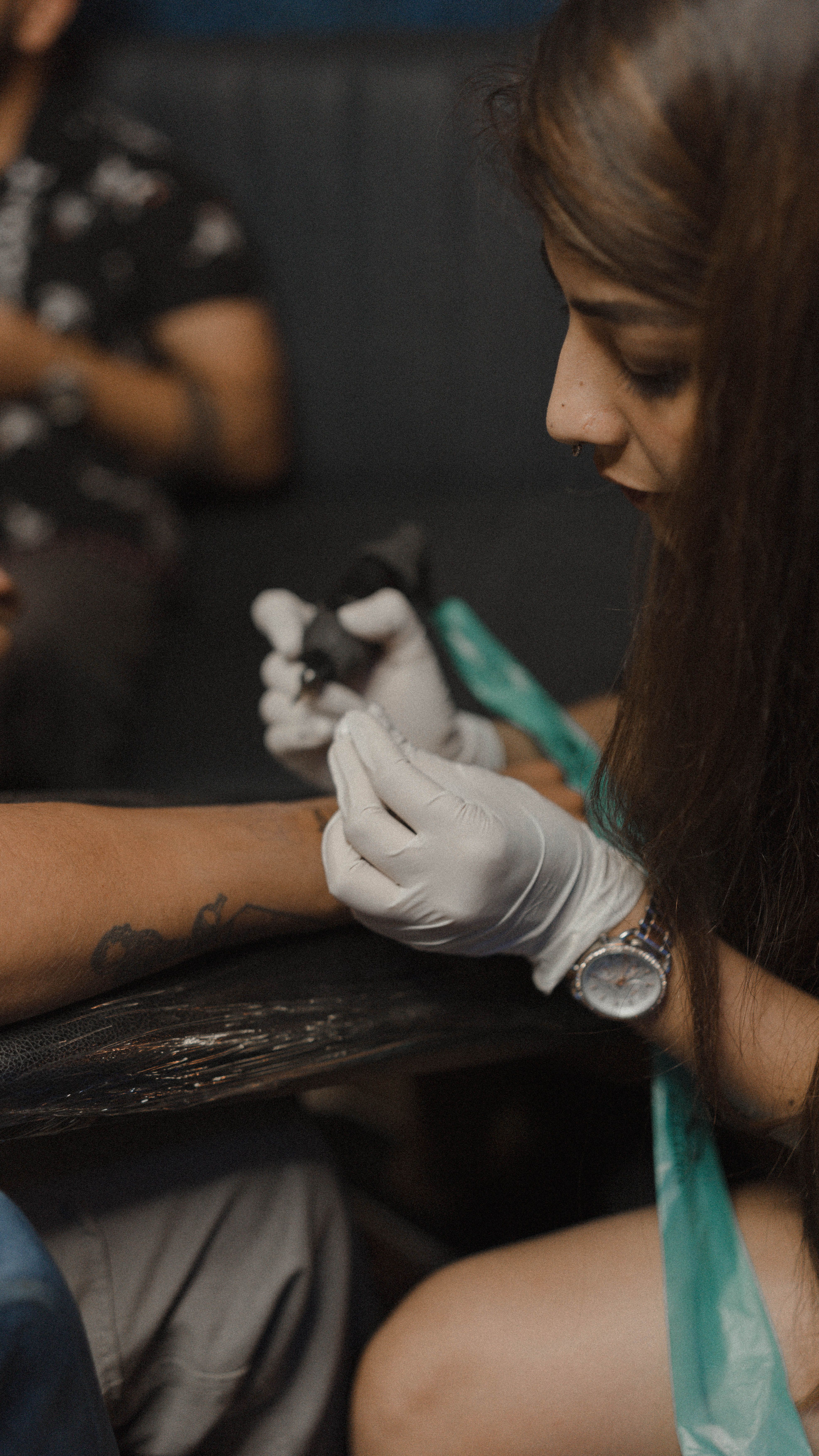 A focused female tattoo artist wearing gloves and a watch, tattooing a client's arm indoors.
