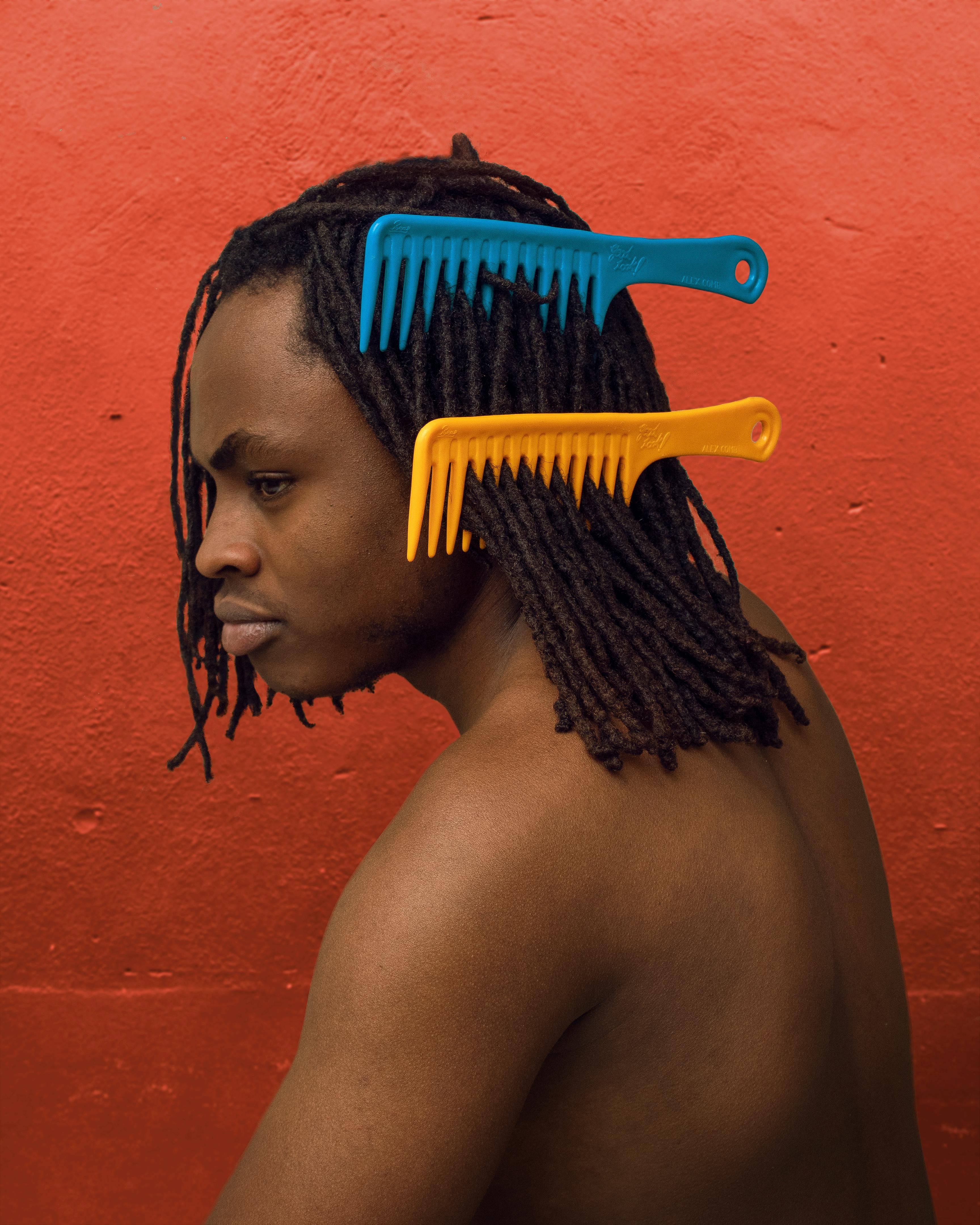Artistic portrait of a shirtless man with dreadlocks and vibrant combs against a red background.