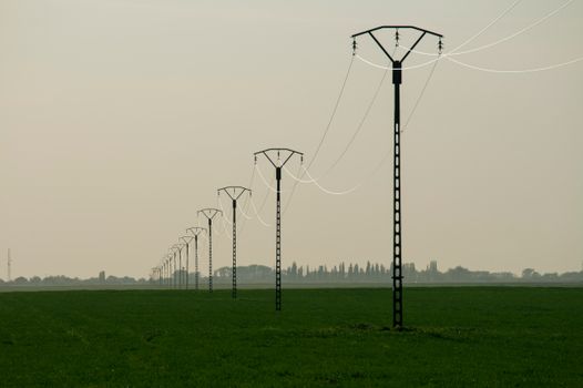 A row of electrical poles across a vast green field under a hazy sky, depicting energy infrastructure.