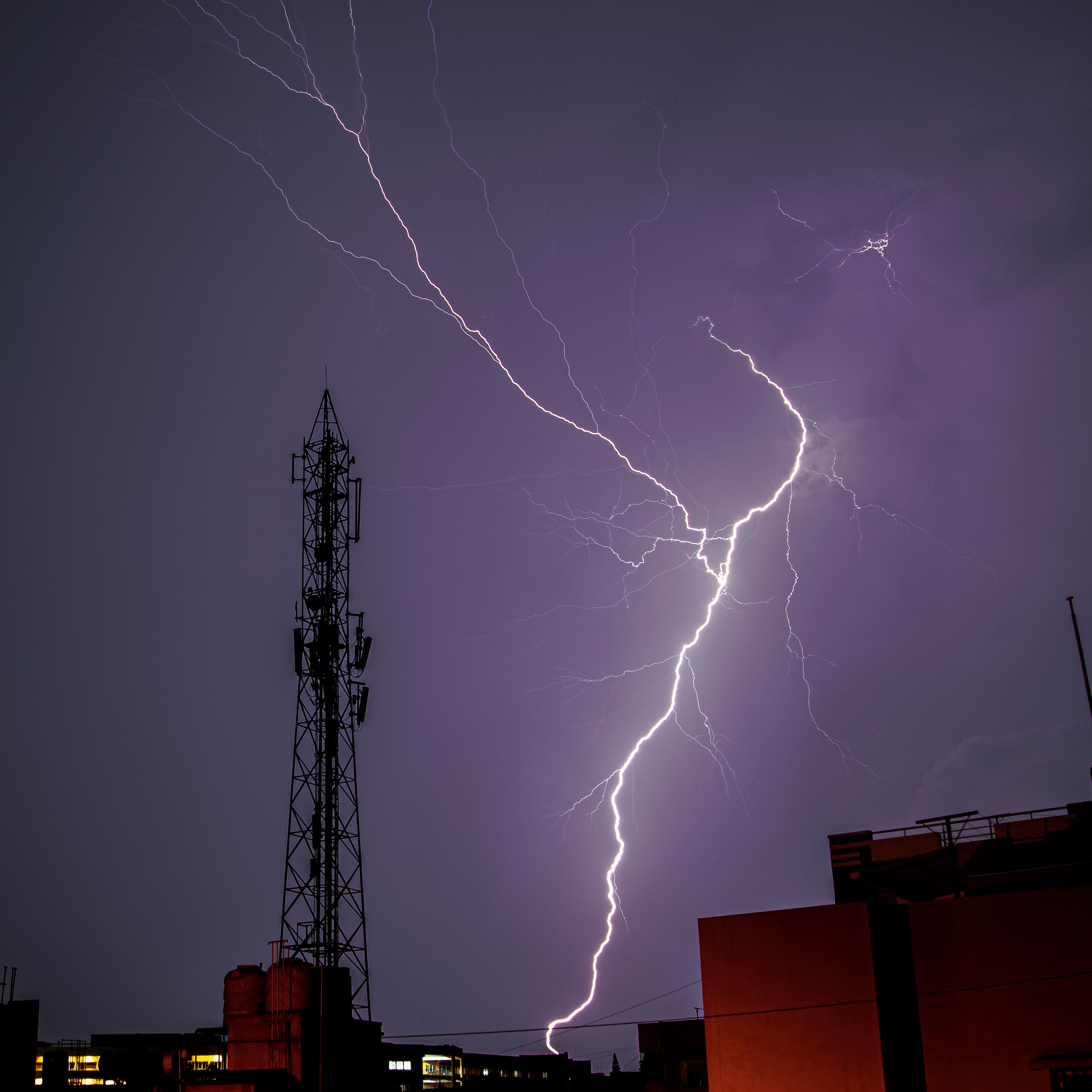 Intense lightning bolt striking near an antenna in a cityscape, captured during a thunderstorm.